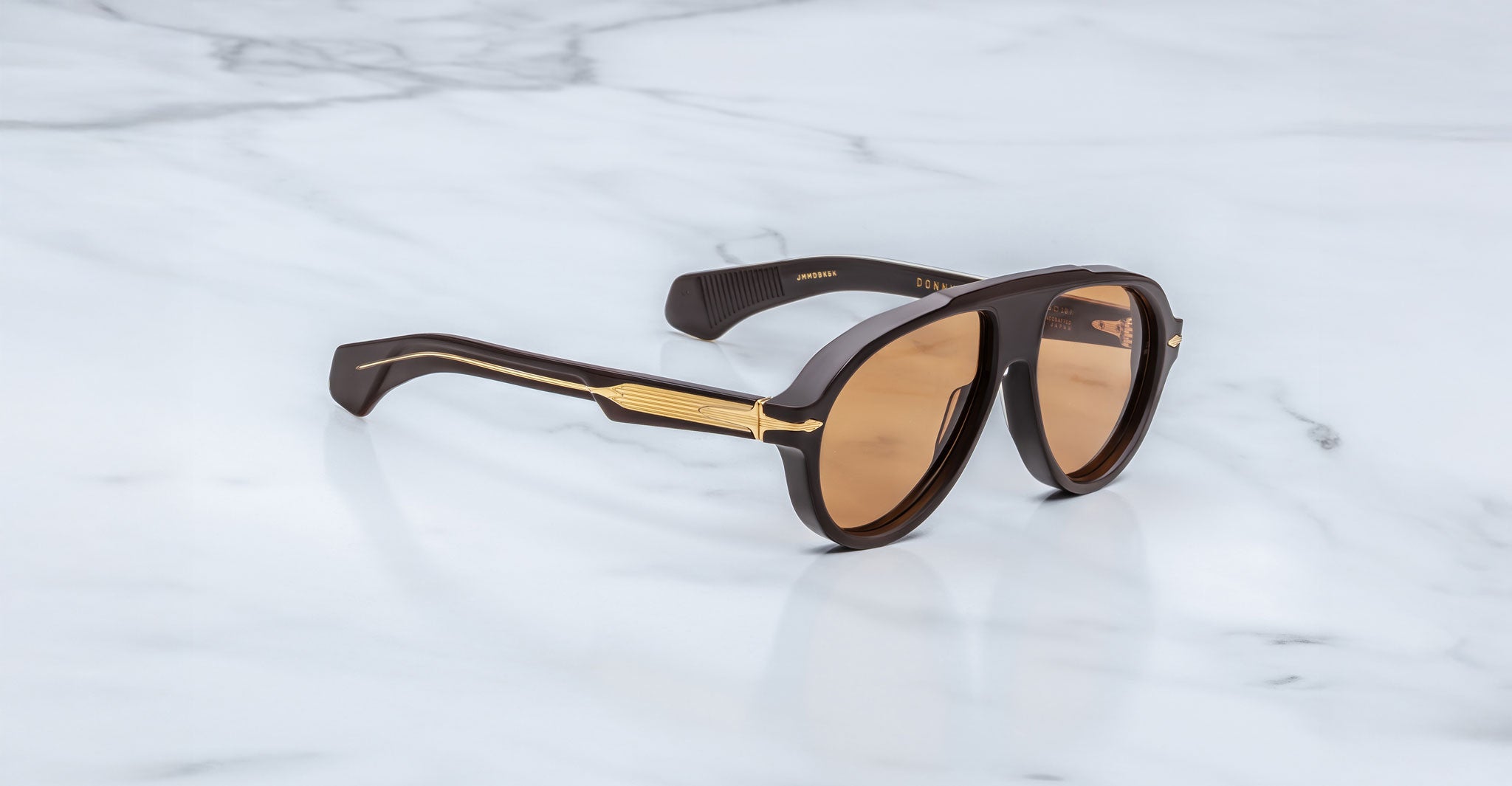 The Jacques Marie Mage Donnybrooke limited-edition sunglasses feature brown oval lenses and gold-accented temples, displayed elegantly on a white marble surface.