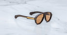 The Jacques Marie Mage Donnybrooke limited-edition sunglasses feature brown oval lenses and gold-accented temples, displayed elegantly on a white marble surface.