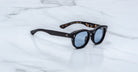 Jacques Marie Mage Dr. Dream sunglasses—black, round frames with blue lenses—are folded on a white marble surface, highlighting the luxury and Japanese craftsmanship of Jacques Marie Mage eyewear.