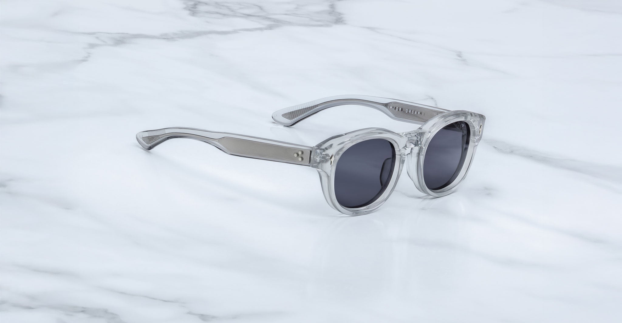 The Dr. Dream sunglasses by Jacques Marie Mage, featuring clear luxury frames and round dark lenses, rest on a white marble surface.