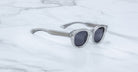 The Dr. Dream sunglasses by Jacques Marie Mage, featuring clear luxury frames and round dark lenses, rest on a white marble surface.