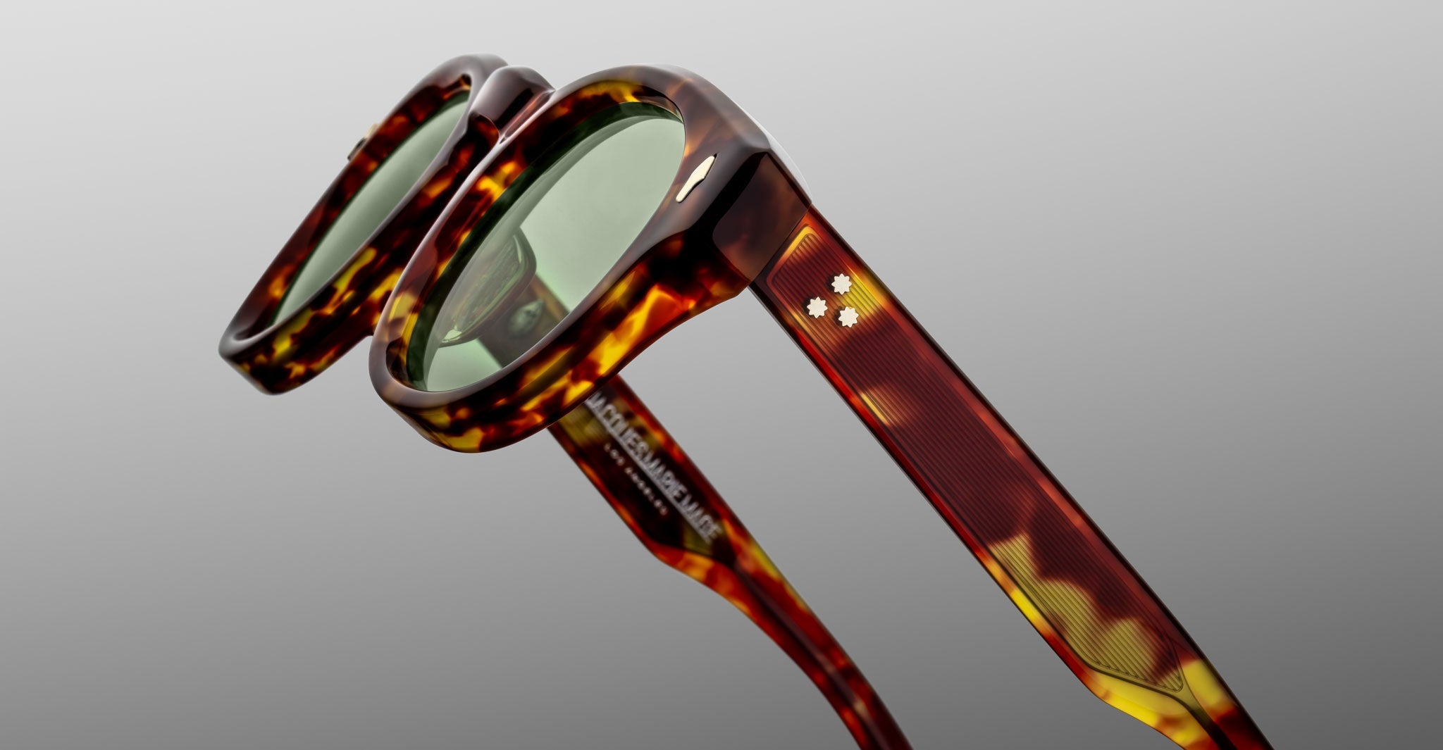 Close-up of Jacques Marie Mage Dr. Dream sunglasses featuring green lenses and two white star-shaped accents on the temples, highlighting luxurious tortoiseshell frames by Jacques Marie Mage against a plain gray background.