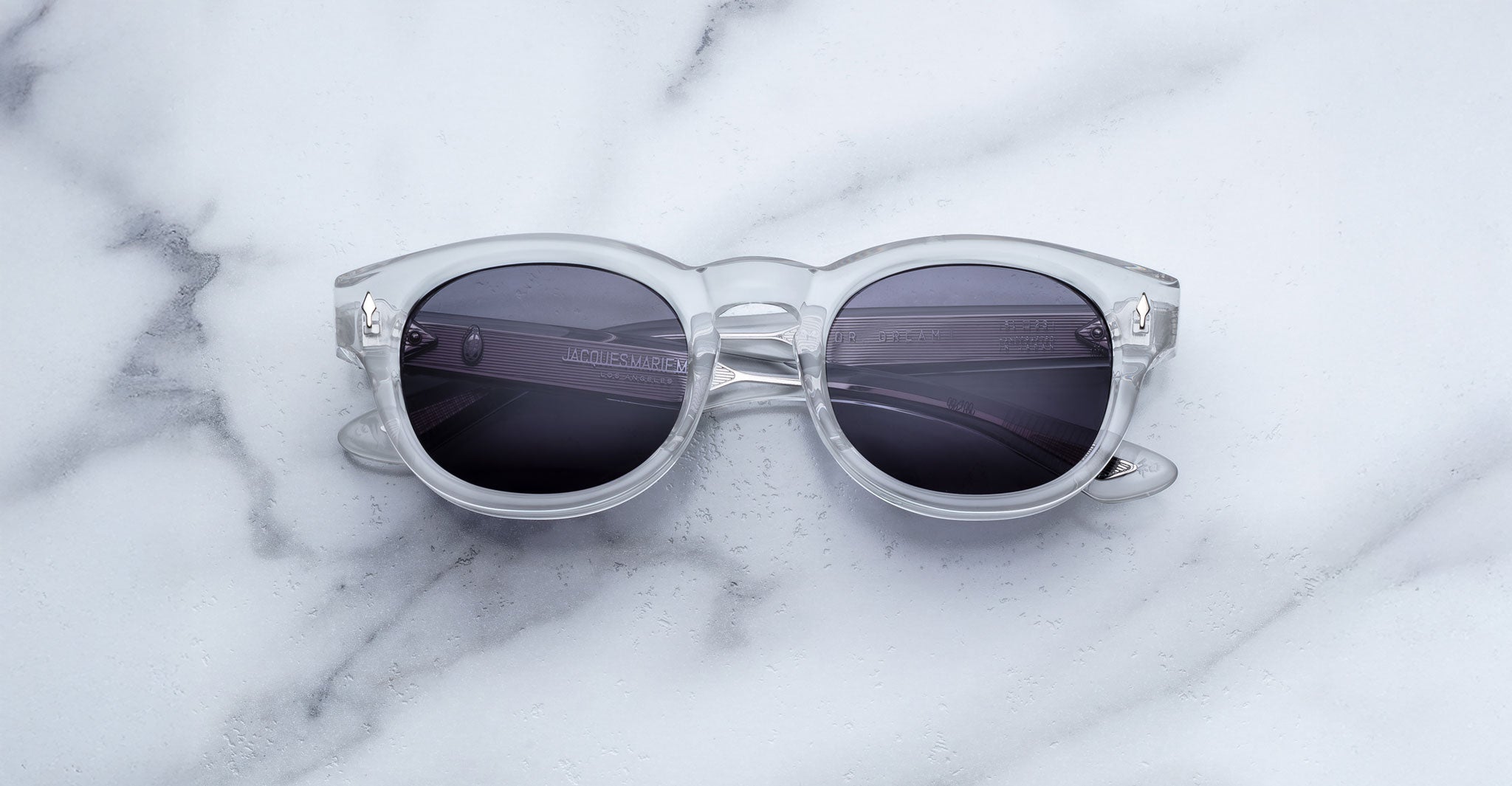The Jacques Marie Mage Dr. Dream sunglasses by Jacques Marie Mage, featuring clear frames and dark lenses, are displayed on a white marble surface.