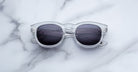 Jacques Marie Mage Dr. Dream sunglasses with clear frames and dark lenses rest on white marble, highlighting the brand’s luxury craftsmanship and refined design.