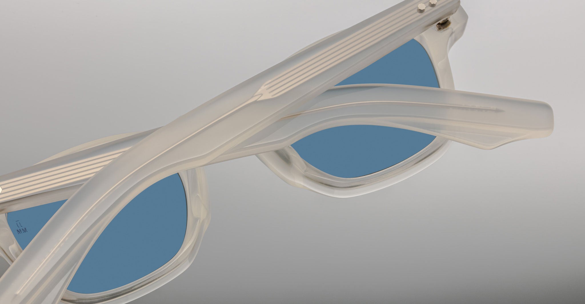 Close-up of Jacques Marie Mage Zephirin sunglasses in translucent white with blue lenses, shown angled with crossed arms—a stunning, limited edition model from Jacques Marie Mage.