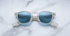 Limited edition Jacques Marie Mage Zephirin sunglasses feature a translucent white acetate frame and blue-tinted lenses, capturing the elegance of handcrafted eyewear. Displayed on a white marble surface by Jacques Marie Mage.