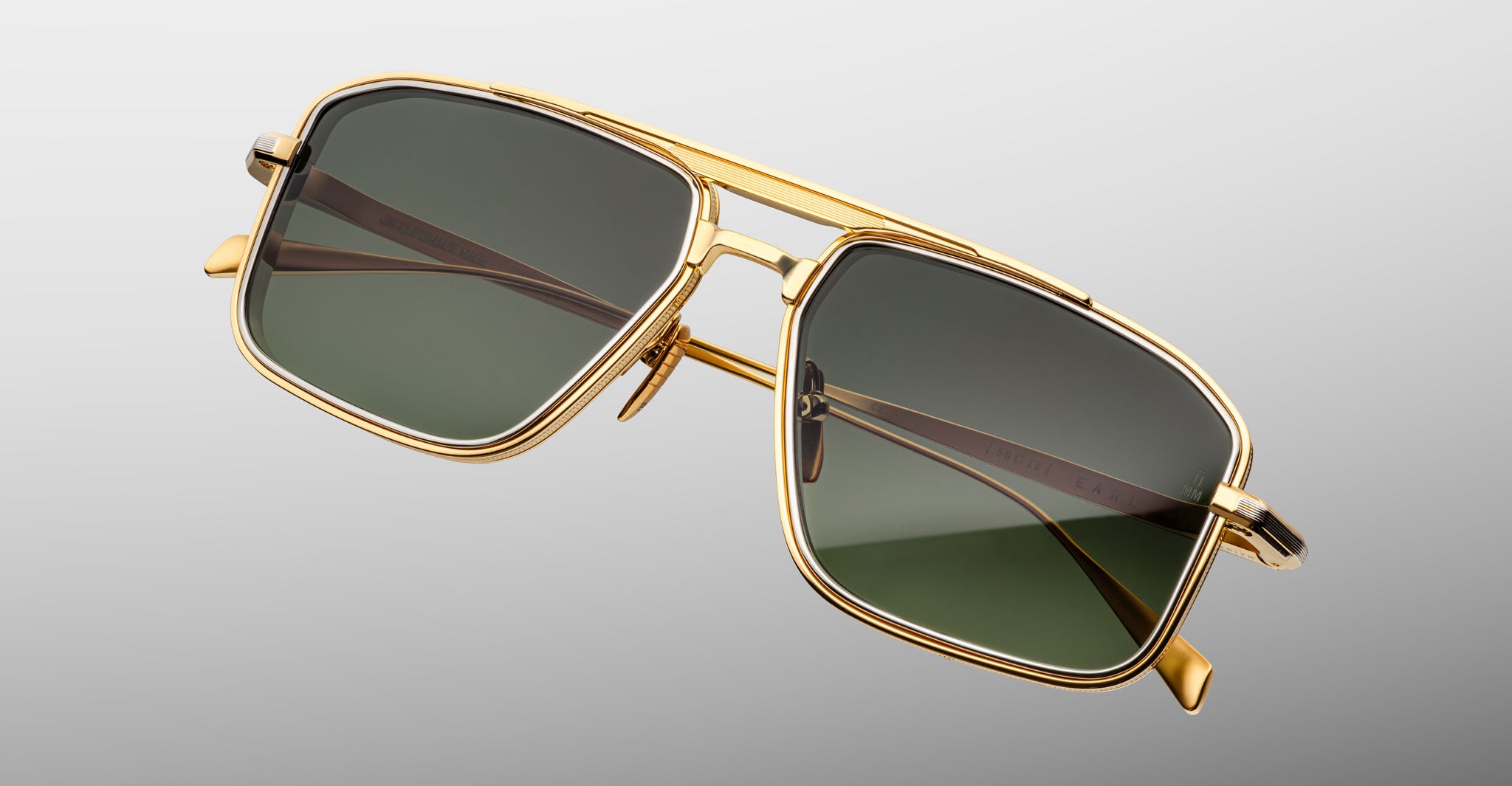 The Jacques Marie Mage Earl sunglasses feature gold aviator frames with green-tinted lenses, a double bridge, and brown arm accents, styled against a light gray background for a ‘70s-inspired look.