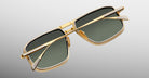 The Jacques Marie Mage Earl sunglasses feature stylish ’70s-inspired gold frames and green-tinted rectangular lenses, displayed from an overhead angle on a light gray gradient background.