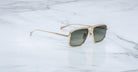Jacques Marie Mage Earl gold-framed aviator sunglasses with dark lenses are displayed on a white marble surface with subtle grey veining.