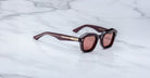 The Jacques Marie Mage Eichler is a pair of dark brown square sunglasses with gold accents on the arms, tinted brown lenses, and thick frames, shown resting on a white marble surface.