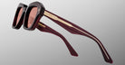 A close-up of Jacques Marie Mage Eichler sunglasses with thick, square frames, dark red lenses, and maroon arms featuring gold detailing and the brand name on the left arm, shown against a grey background.