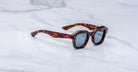 Jacques Marie Mage Eichler sunglasses with a tortoiseshell pattern, thick angular frames, and dark lenses are displayed on a white marble surface featuring subtle grey veining.