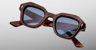 The Jacques Marie Mage Eichler sunglasses, featuring thick angular tortoiseshell frames and blue-tinted lenses, are displayed against a plain light gray background.