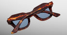 The Jacques Marie Mage Eichler sunglasses feature a tortoiseshell frame with blue lenses, displayed folded and viewed from the back on a light gray background.