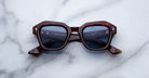 The Jacques Marie Mage Eichler sunglasses feature a brown tortoiseshell hexagonal frame with blue lenses, displayed on a white marble surface.