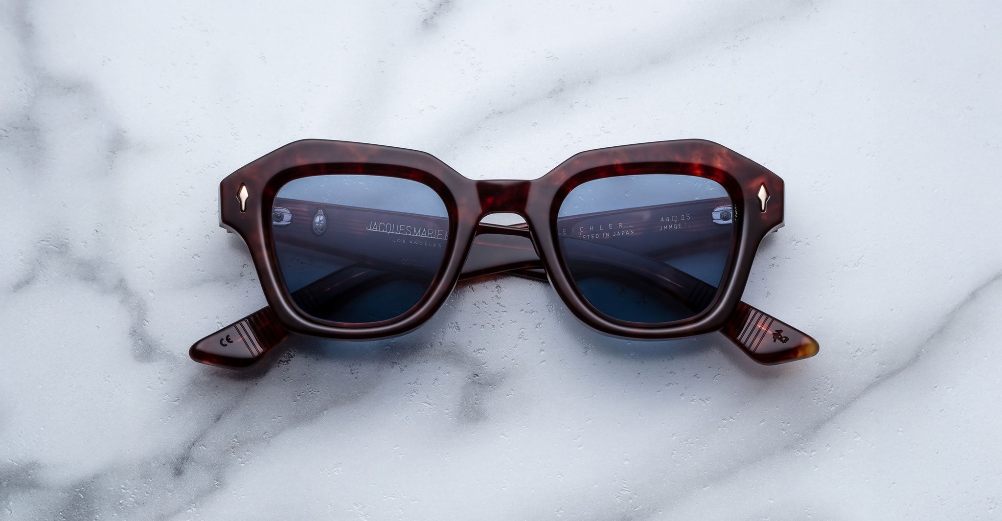 The Jacques Marie Mage Eichler sunglasses feature a brown tortoiseshell hexagonal frame with blue lenses, displayed on a white marble surface.