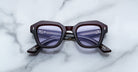 A pair of Jacques Marie Mage Eichler cherry color, thick-framed sunglasses with lilac lenses rests on a white marble surface with grey veins, with faint brand details visible on the inside of the left lens.