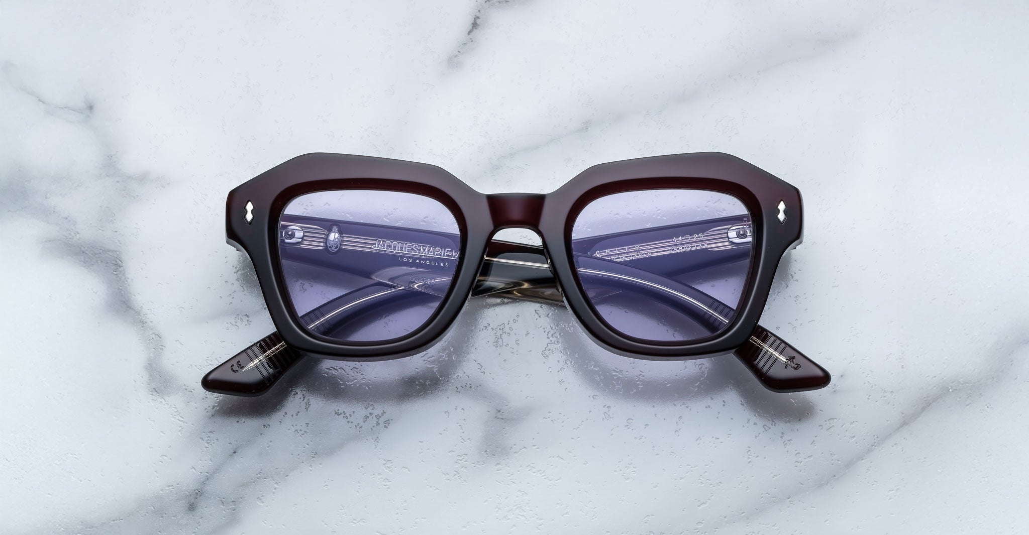 A pair of Jacques Marie Mage Eichler cherry color, thick-framed sunglasses with lilac lenses rests on a white marble surface with grey veins, with faint brand details visible on the inside of the left lens.