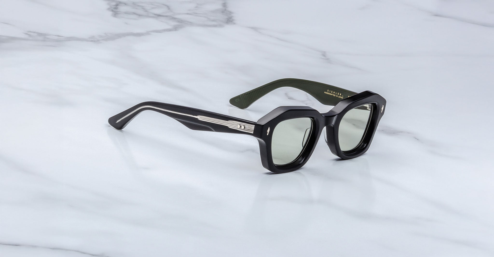 A pair of Jacques Marie Mage Eichler sunglasses, featuring black square frames, silver temple detailing, and green-tinted lenses, rests on a white marble surface.