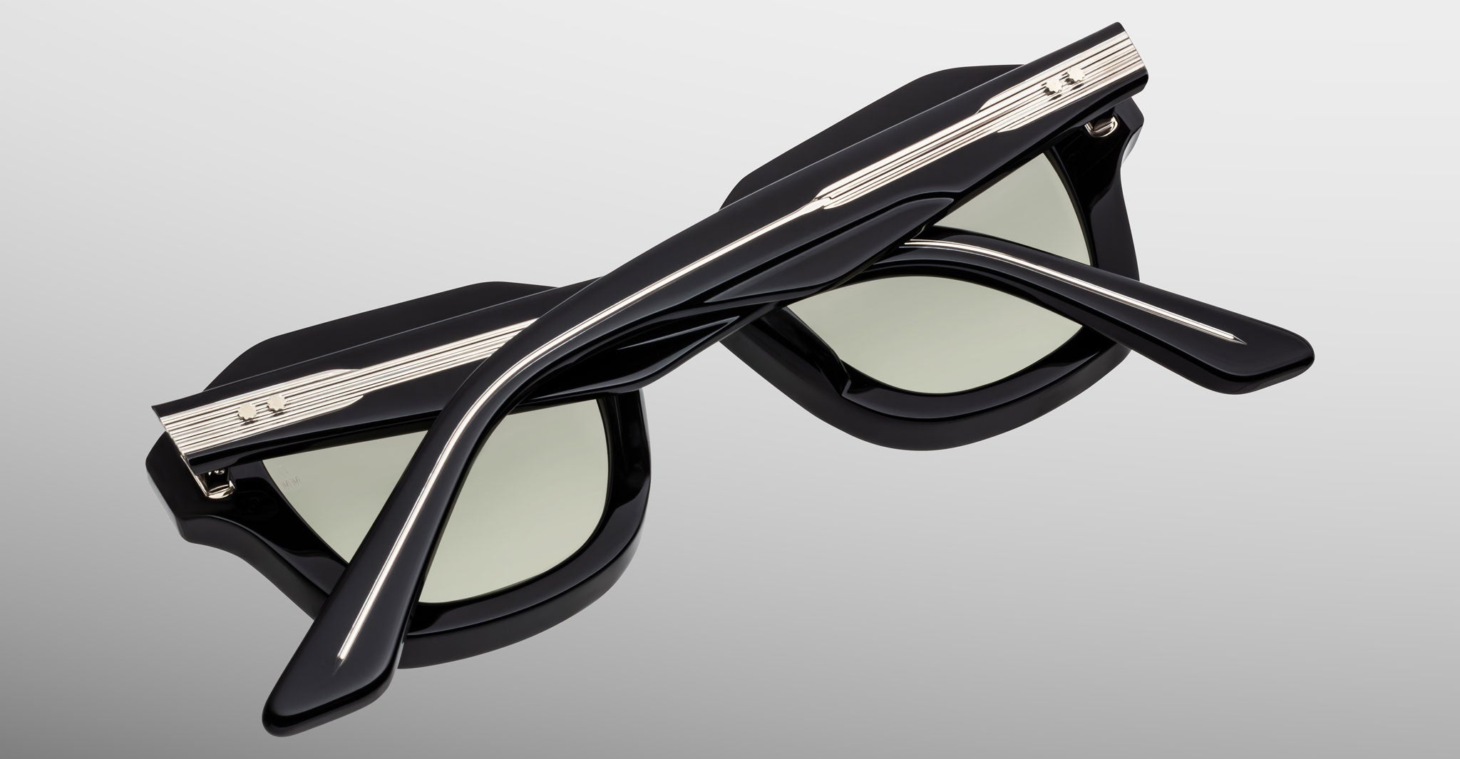 The Jacques Marie Mage Eichler sunglasses are shown upside down against a gray background, showcasing their black frames, light-tinted lenses, and distinctive metallic detailing on the arms and top of the frame.