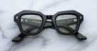 A pair of Jacques Marie Mage Eichler thick black rectangular eyeglasses with green-tinted lenses rests on a white marble surface. Gold text on the inside arms details the brand and model.