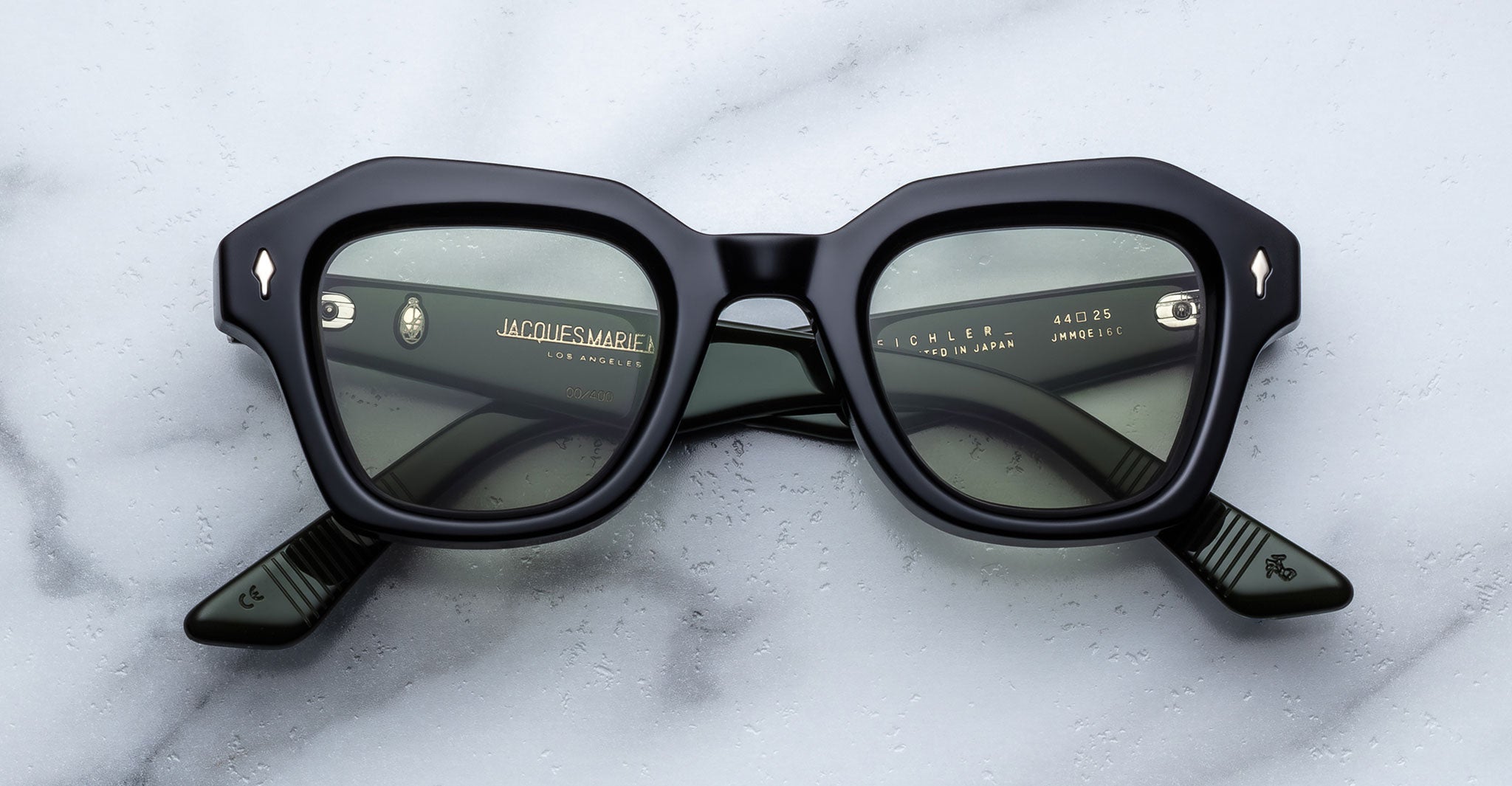 A pair of Jacques Marie Mage Eichler thick black rectangular eyeglasses with green-tinted lenses rests on a white marble surface. Gold text on the inside arms details the brand and model.