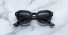 A pair of Jacques Marie Mage Eichler black, thick-framed sunglasses with grey lenses rests on a white marble surface with grey veins, with faint brand details visible on the inside of the left lens.
