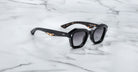 A pair of Jacques Marie Mage Eichler black, thick-framed sunglasses with grey lenses sits on a white marble surface with grey veins