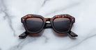 A pair of Jacques Marie Mage Eichler Havana color, thick-framed sunglasses with brown lenses rests on a white marble surface with grey veins, with faint brand details visible on the inside of the left lens.