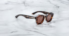 A pair of Jacques Marie Mage Eichler Havana, thick-framed sunglasses with brown lenses sits on a white marble surface with grey veins