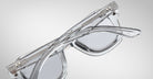 A pair of Jacques Marie Mage Eichler crystal, thick-framed sunglasses with light grey lenses rests on a white marble surface with grey veins, with faint brand details visible on the inside of the left lens.