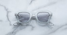 A pair of Jacques Marie Mage Eichler crystal, thick-framed sunglasses with grey lenses rests on a white marble surface with light grey veins, with faint brand details visible on the inside of the left lens.