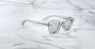 Jacques Marie Mage Eichler sunglasses, featuring clear geometric frames and silver-tinted lenses, rest on a white marble surface with gray veining.