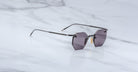 The Jacques Marie Mage El Dorado features geometric rimless sunglasses with dark tinted lenses and slim metal arms, displayed on a light gray marble surface.