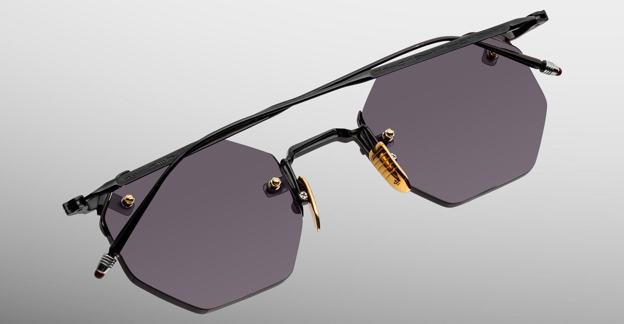 The Jacques Marie Mage El Dorado: sleek sunglasses featuring dark hexagonal lenses, slim black metal frames, gold nose pads, and a minimalist design, displayed on a light gray gradient background.