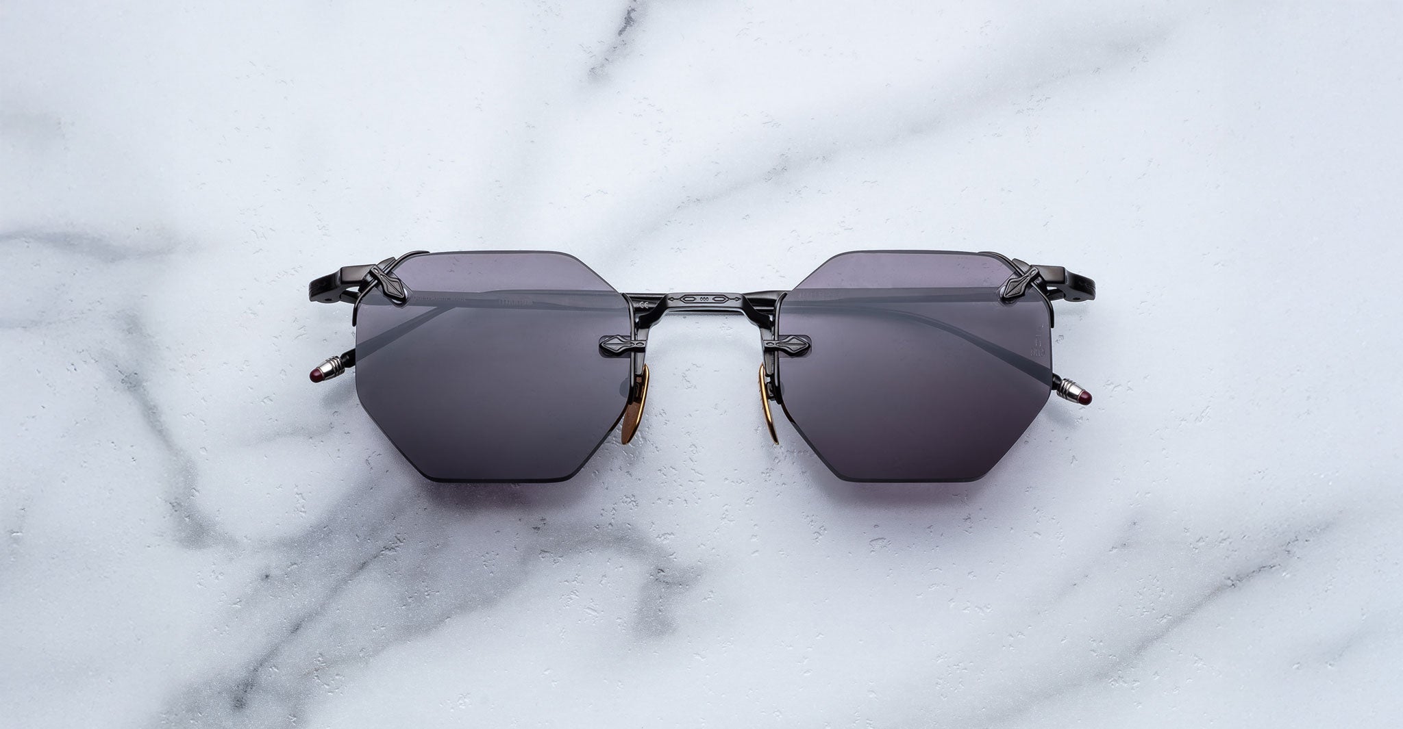 The Jacques Marie Mage El Dorado sunglasses, featuring geometric hexagonal black frames and dark lenses, are displayed on a white marble surface.
