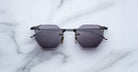 The Jacques Marie Mage El Dorado sunglasses, featuring geometric hexagonal black frames and dark lenses, are displayed on a white marble surface.