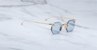 The Jacques Marie Mage El Dorado features octagonal, rimless sunglasses with blue tinted lenses and slim gold metal arms, displayed on a white marble surface.