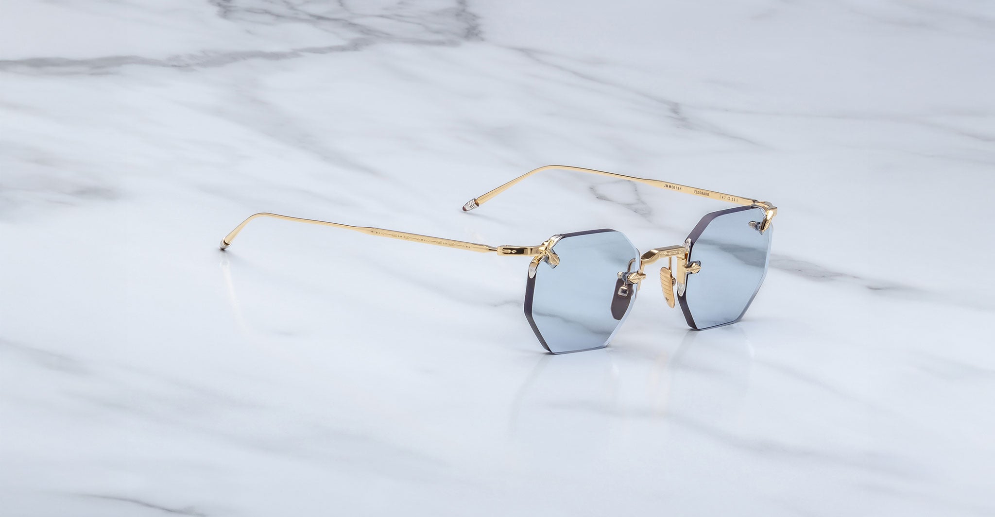 The Jacques Marie Mage El Dorado features octagonal, rimless sunglasses with blue tinted lenses and slim gold metal arms, displayed on a white marble surface.