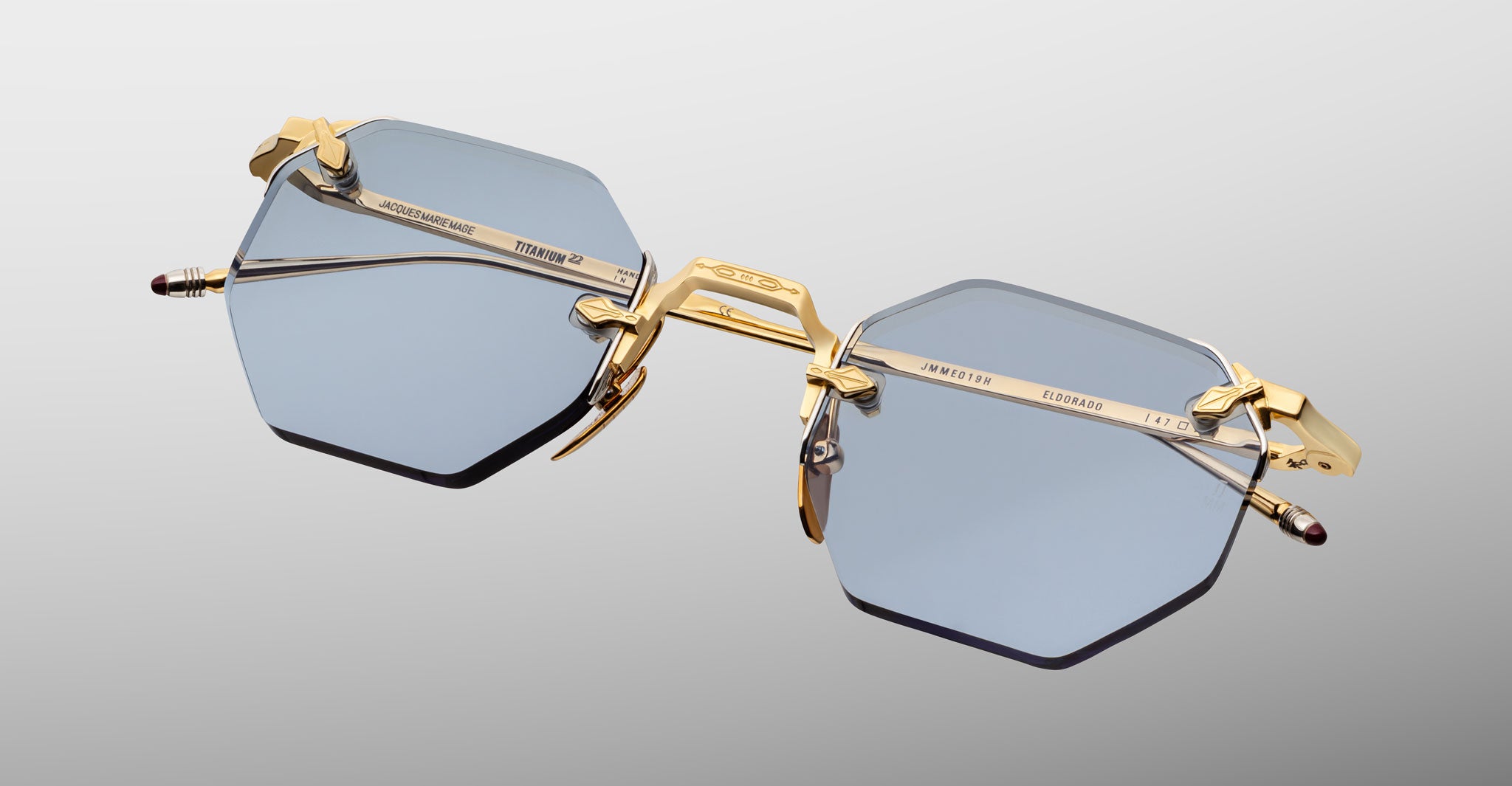 The Jacques Marie Mage El Dorado features rimless hexagonal blue-tinted lenses with gold metal temples and bridge, showcased against a neutral gradient background.
