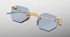 The Jacques Marie Mage El Dorado features rimless hexagonal blue-tinted lenses with gold metal temples and bridge, showcased against a neutral gradient background.