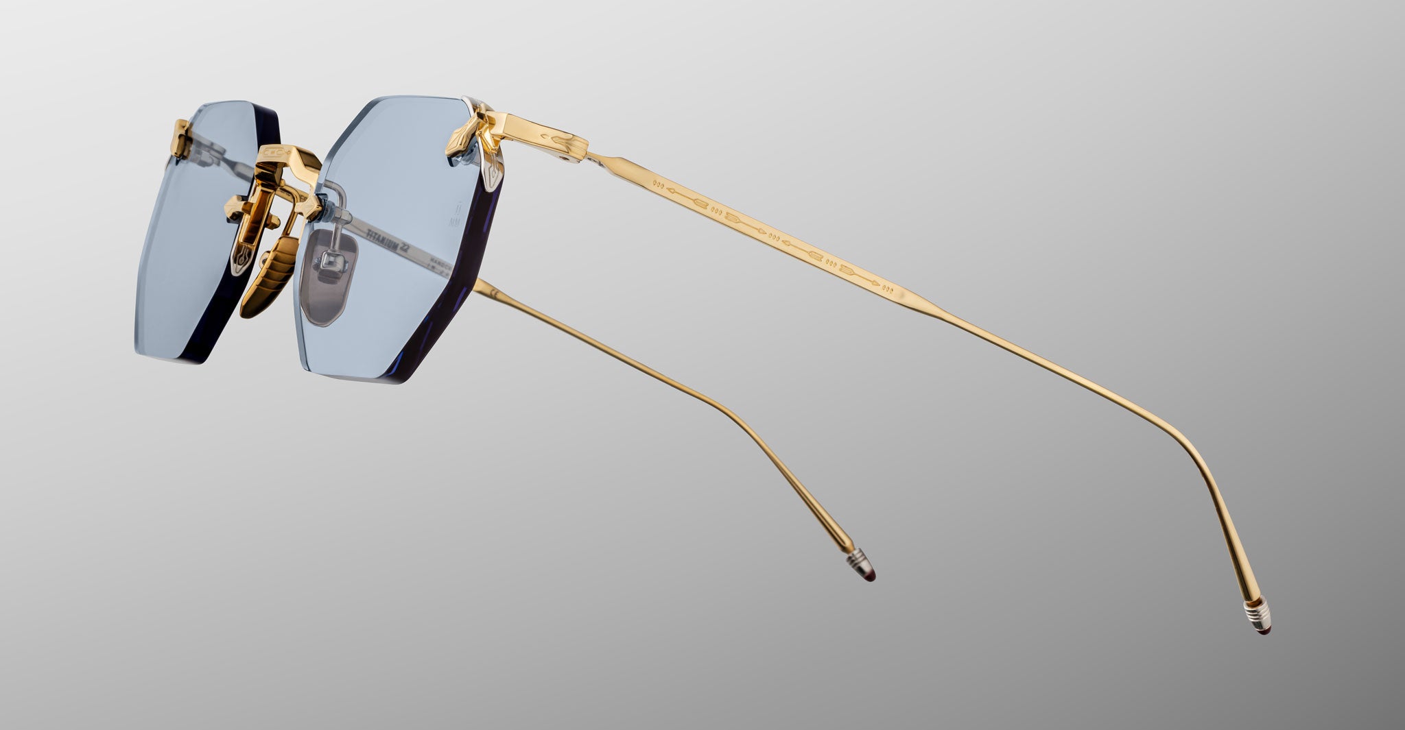 The Jacques Marie Mage El Dorado features rimless eyeglasses with blue-tinted rectangular lenses and slim gold metal arms, displayed against a light gray gradient background.