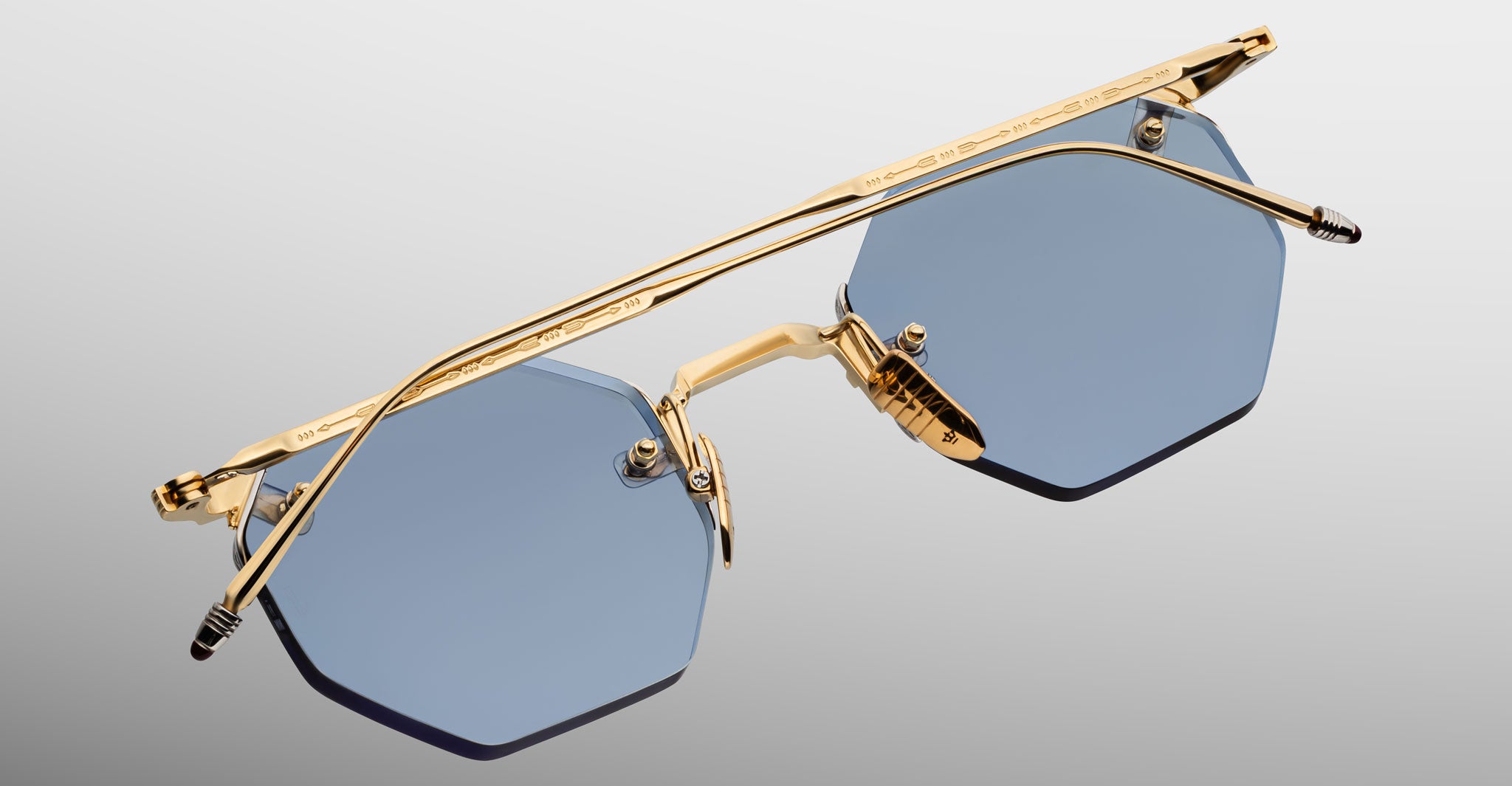 The Jacques Marie Mage El Dorado sunglasses feature a geometric design with blue-tinted lenses and a gold metal frame, showcased from above against a light gradient background.