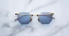 The Jacques Marie Mage El Dorado sunglasses, featuring hexagonal blue lenses and gold metal frames, are displayed on a white marble surface.