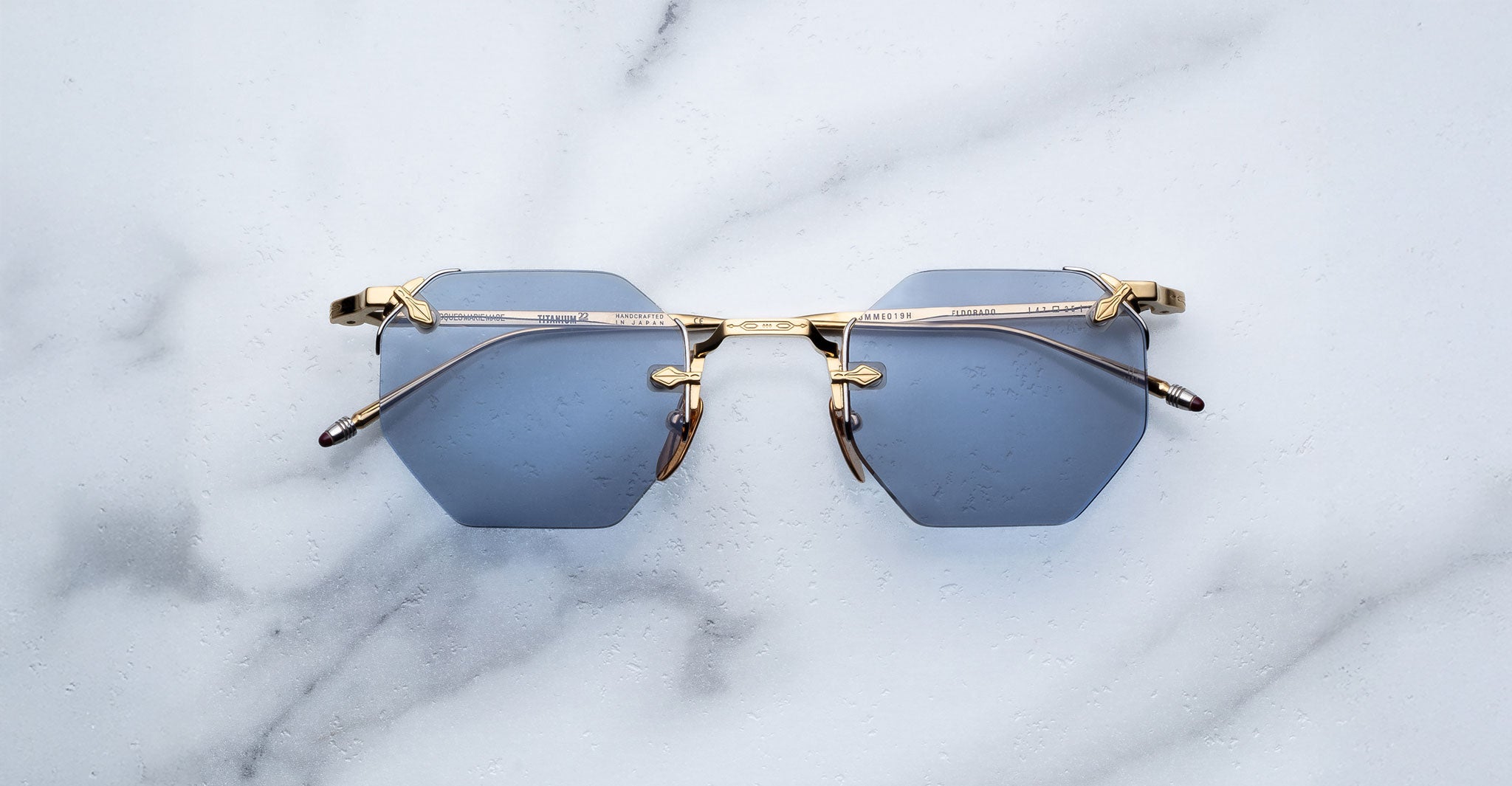 The Jacques Marie Mage El Dorado sunglasses, featuring hexagonal blue lenses and gold metal frames, are displayed on a white marble surface.