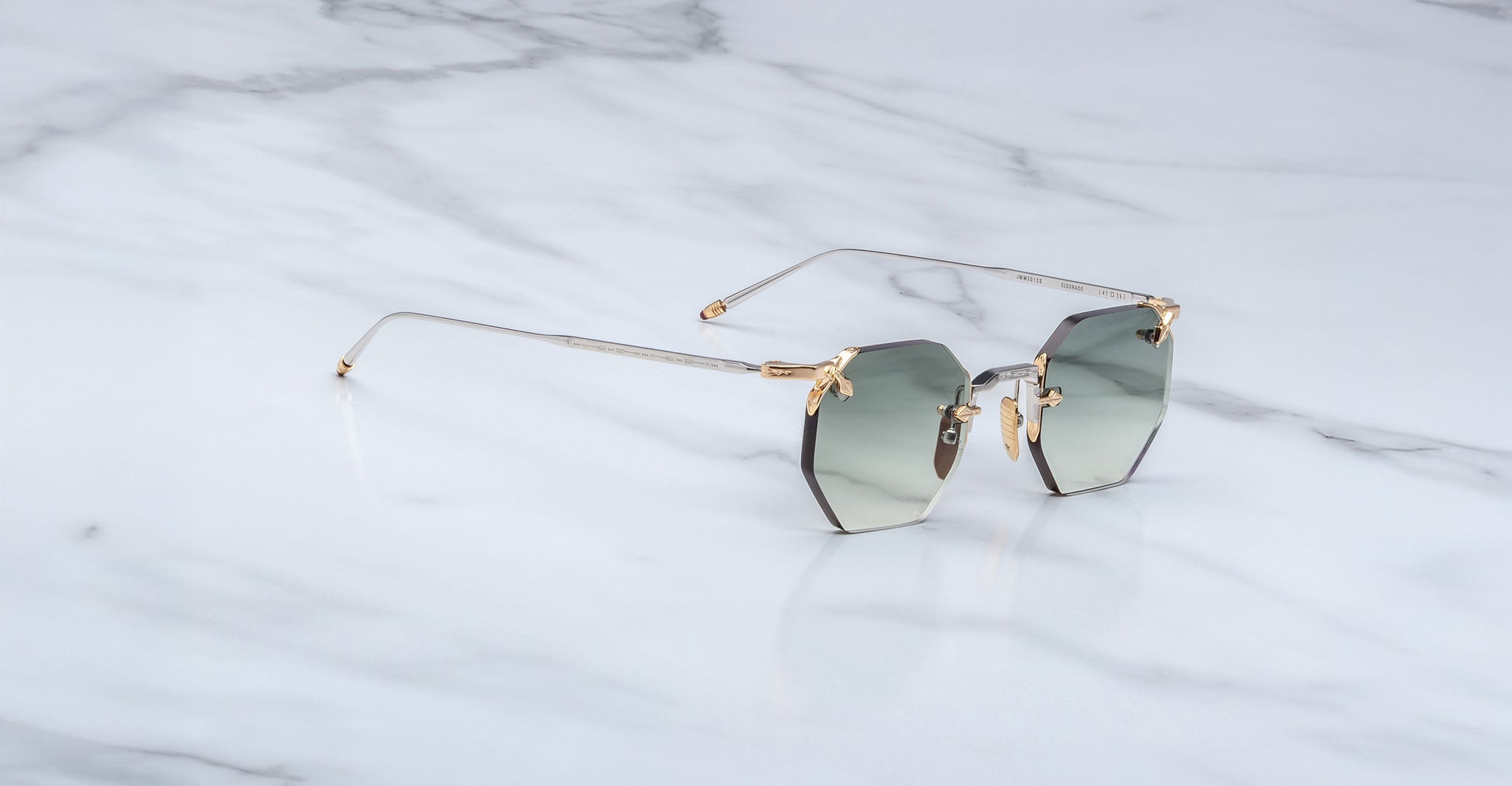A pair of Jacques Marie Mage El Dorado geometric rimless sunglasses with gray gradient lenses and thin gold-toned arms rests on a white marble surface.