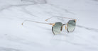 A pair of Jacques Marie Mage El Dorado geometric rimless sunglasses with gray gradient lenses and thin gold-toned arms rests on a white marble surface.