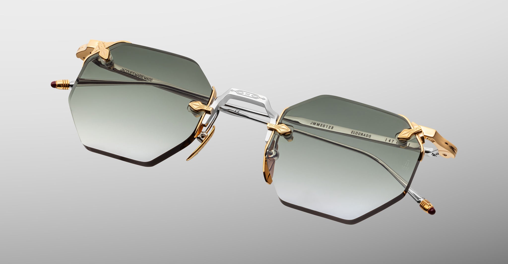 The Jacques Marie Mage El Dorado features hexagonal sunglasses with gradient gray lenses, gold and silver metal frames, and gold accents on the hinges and nose pads, shown against a light gray background.