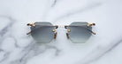 The Jacques Marie Mage El Dorado sunglasses, featuring geometric hexagonal frames with gray lenses and gold accents, are displayed on a white marble surface.