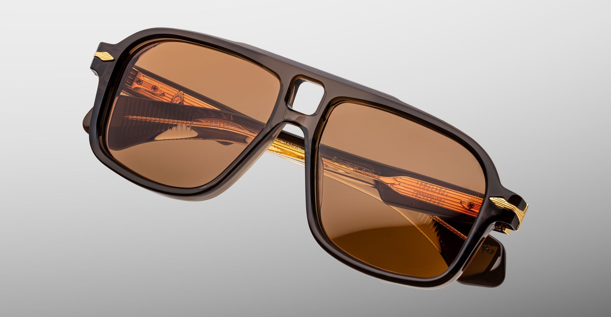 The Jacques Marie Mage Elkhart sunglasses feature a brown aviator-style frame, amber-tinted lenses, gold accents, and a sculptural brow ridge, displayed on a grey background. Inspired by the brand's limited-edition navigator designs.
